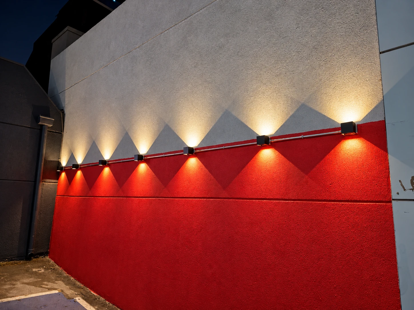 Siete apliques encendidos proyectan luz hacia arriba y abajo sobre un muro rojo y gris